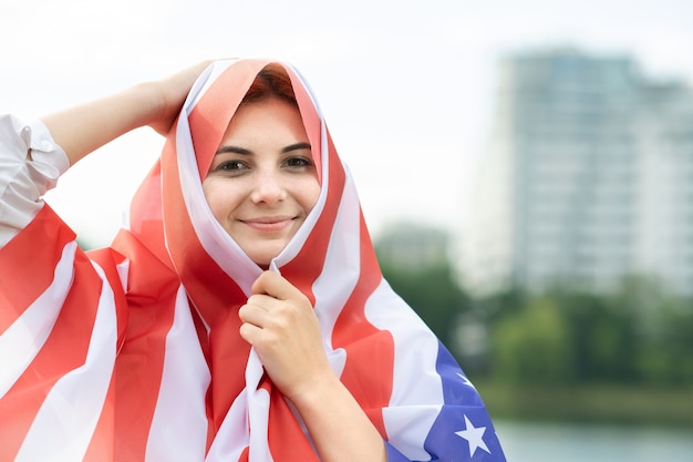 Portret van jonge gelukkige vluchtelingvrouw met de nationale vlag van de V.S. op haar hoofd en schouders. Positief moslimmeisje dat de onafhankelijkheidsdag van de Verenigde Staten viert.