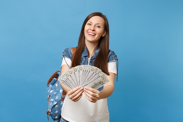Portret van jonge glimlachende aantrekkelijke vrouw student in denim kleding met rugzak met bundel veel dollars, contant geld geïsoleerd op blauwe achtergrond. Onderwijs in middelbare school hogeschool.