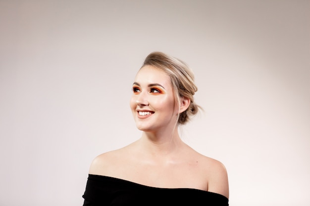 Portret van jonge glimlachende blondevrouw met professionele make-up. Vrouw in zwarte trui met blote schouders poseren