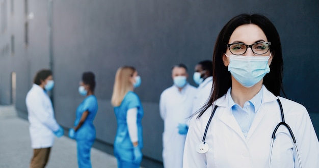 Portret van jonge Kaukasische brunette mooie vrouw arts in medisch masker camera kijken. Close up van vrouwelijke arts in ademhalingsbescherming. Medics-collega's van gemengde rassen op achtergrond.