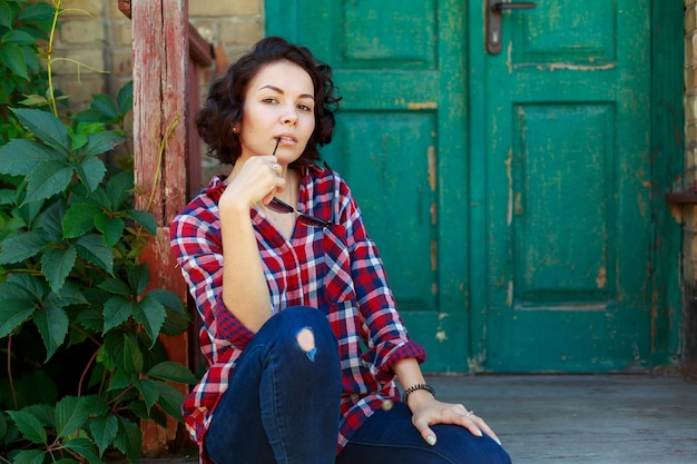 Portret van jonge krullende vrouw dichtbij oude huismuur. Glimlachend en emotioneel meisje in jeans en rood shirt en zonnebril op de stadsstraat. Leuke jonge vrouw in openlucht in zonnige dag