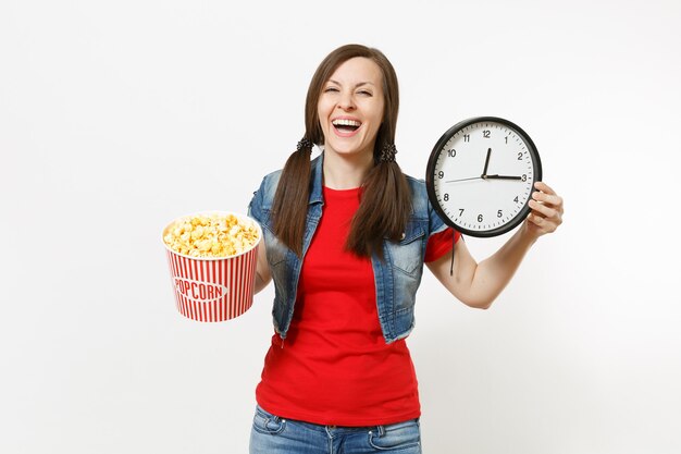 Portret van jonge lachende aantrekkelijke brunette vrouw in casual kleding kijken naar film, emmer popcorn en ronde wekker geïsoleerd op een witte achtergrond. Emoties in bioscoopconcept.