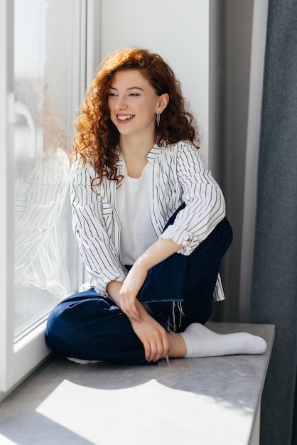 Portret van jonge lachende roodharige vrouw zittend op de vensterbank