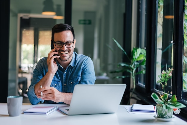 Portret van jonge lachende vrolijke ondernemer in casual kantoor bellen tijdens het werken met laptop