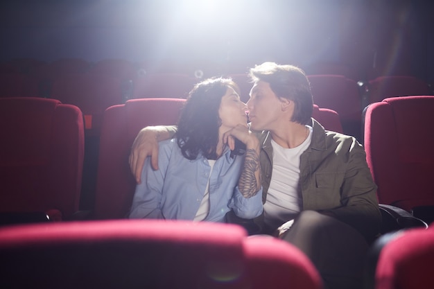 Portret van jonge liefdevolle paar zoenen in de bioscoop terwijl u geniet van datum, man omhelst vriendin in donkere zaal, kopie ruimte