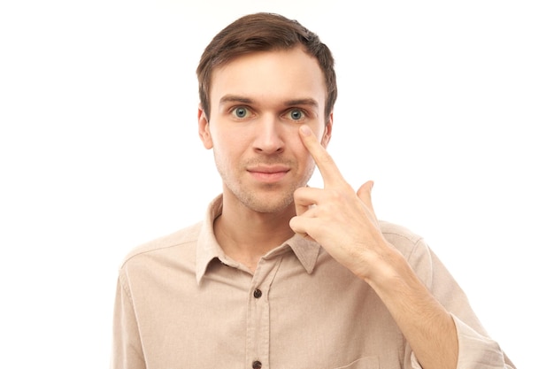 Portret van jonge man wijst met de vinger naar zijn oog geïsoleerd op witte studio achtergrond Visiecontrole en verlies van gezichtsvermogen concept brillenwinkel