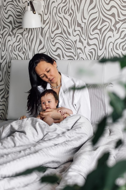 Portret van jonge moeder en mooie dochter in slaapkamer wazig groene planten op voorgrond gratis kopieerruimte Rusten in grijs bed met naakte baby Concept van moederlijke genegenheid en kinderopvang