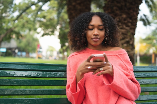 Portret van jonge mooie afro Amerikaanse vrouw met behulp van haar mobiele telefoon en zittend op een bankje in een park. Buitenshuis.