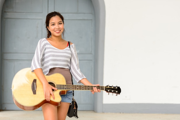 Portret van jonge mooie Aziatische vrouw gitaarspelen in de straten van de stad buitenshuis