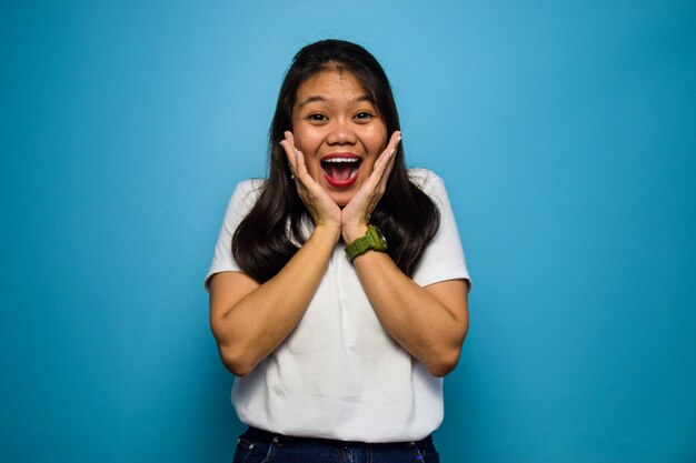 Portret van jonge mooie Aziatische vrouwen die witte T-shirt gebruiken die op blauw wow-gebaar wordt geïsoleerd