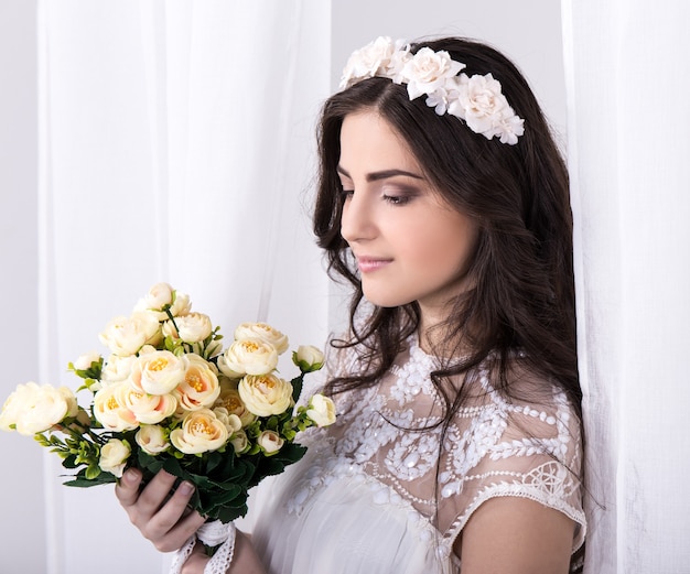 Portret van jonge mooie dromende bruid in witte jurk met bloemen bij het raam