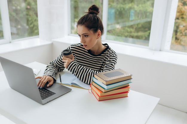 Portret van jonge mooie glimlachende jonge vrouw die aan het bureau zit en geniet van haar kopje koffie terwijl ze werkt of studeert op een laptopcomputer in een klein kantoor aan huis of in het studentenhuis binnenshuis