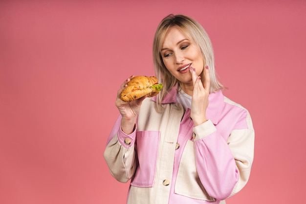 Portret van jonge mooie hongerige vrouw die croissantsandwich eet. Geïsoleerd portret van vrouw met fast food over roze achtergrond. Dieet concept.