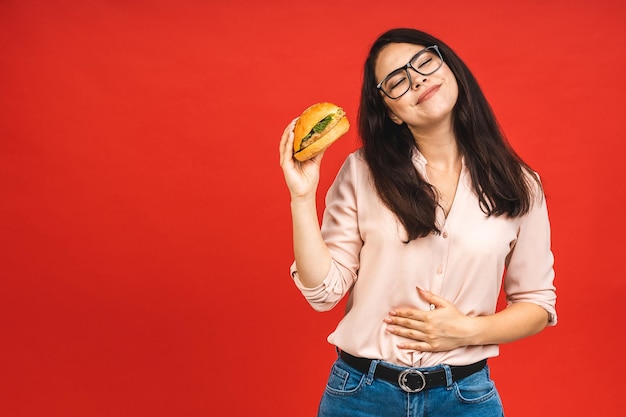 Portret van jonge mooie hongerige vrouw die hamburger eet Geïsoleerd portret van student met fastfood op rode achtergrond Dieetconcept