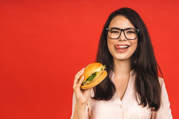 Portret van jonge mooie hongerige vrouw die hamburger eet geïsoleerd portret van student met fastfood op rode achtergrond dieetconcept