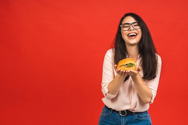 Portret van jonge mooie hongerige vrouw die hamburger eet Geïsoleerd portret van student met fastfood op rode achtergrond Dieetconcept