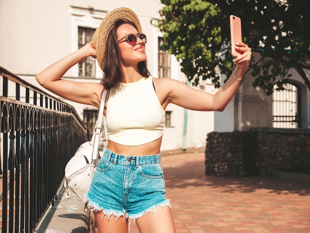 Portret van jonge mooie lachende hipster vrouw in trendy zomer jeans broek en hoed Sexy model poseren op de straat achtergrond bij zonsondergang Positief model buitenshuis met aktetas Selfie nemen