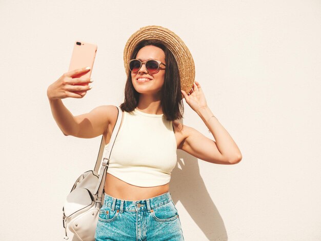 Portret van jonge mooie lachende hipster vrouw in trendy zomer jeans broek en hoed Sexy zorgeloos model poseren in de straat in de buurt van witte muur Positief model buitenshuis met werkmap Selfie nemen