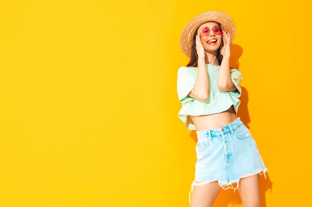 Foto portret van jonge mooie lachende vrouw in trendy zomer jeans rok zorgeloze vrouw poseren in de buurt van gele muur in studio positief model plezier binnenshuis in zonnebril vrolijk en gelukkig in hoed