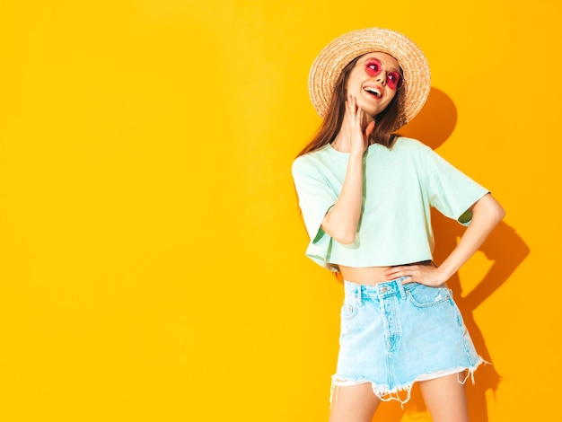 Portret van jonge mooie lachende vrouw in trendy zomer jeans rok zorgeloze vrouw poseren in de buurt van gele muur in studio Positief model plezier binnenshuis vrolijk en gelukkig In hoed