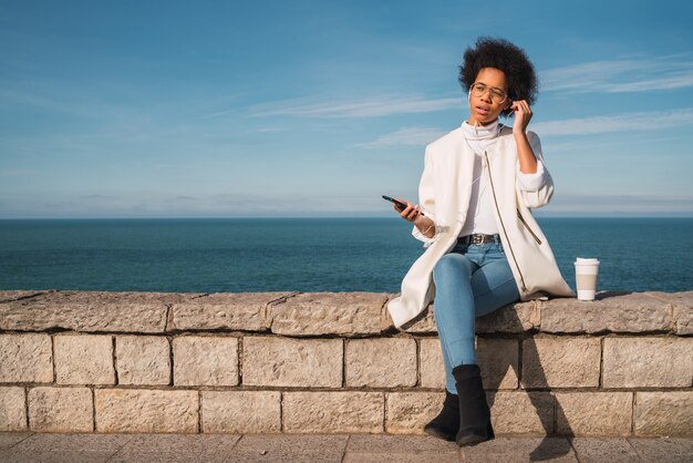 Portret van jonge mooie Latijns-vrouw met behulp van haar mobiele telefoon met koptelefoon, met de zee in de ruimte. Communicatie en technologieconcept.