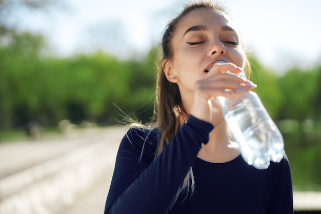 Portret van jonge mooie vrouw die blauw sportkleding drinkwater draagt bij park