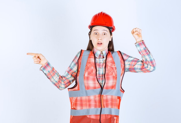 Portret van jonge mooie vrouwelijke ingenieur in helm die ergens naar wijst.