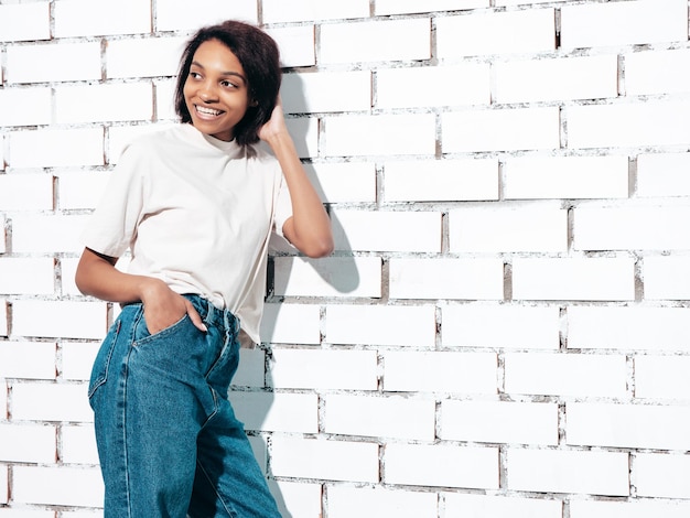 Portret van jonge mooie zwarte vrouw glimlachend model gekleed in zomer jeans en topkleren Sexy zorgeloze vrouw poseren in de buurt van witte bakstenen muur in studio gelooid en vrolijk