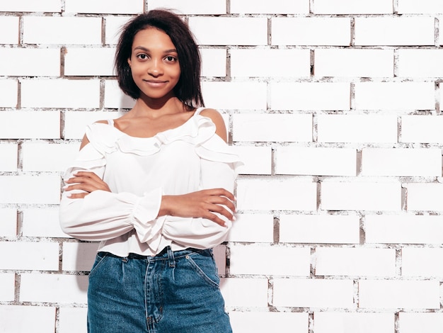 Portret van jonge mooie zwarte vrouw glimlachend model gekleed in zomer jeans kleding Sexy zorgeloze vrouw poseren in de buurt van witte bakstenen muur in studio gelooid en vrolijk