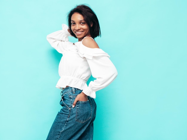 Portret van jonge mooie zwarte vrouw Glimlachend model gekleed in zomerse jeans kleding Sexy zorgeloze vrouw poseren in de buurt van blauwe muur in studio Gelooid en vrolijk