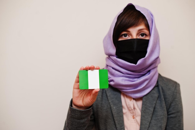 Portret van jonge moslimvrouw die formele kleding draagt, beschermt gezichtsmasker en hijab-hoofddoek houdt Nigeriaanse vlagkaart vast tegen geïsoleerde achtergrond Coronavirus landconcept