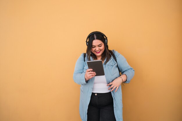 Portret van jonge plus groottevrouw die aan muziek met hoofdtelefoons en digitale tablet in openlucht tegen gele muur luistert.