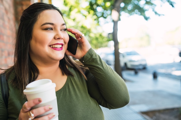 Portret van jonge plus size vrouw praten aan de telefoon terwijl ze een kopje koffie buiten op straat houdt