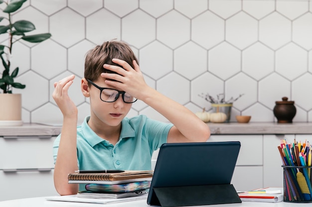 Portret van jonge preteen overstuur jongen kijkend naar notebook in de buurt van tablet die voorhoofd aanraakt met hand die online les heeft