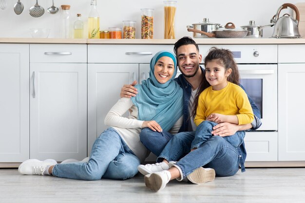 Portret van jonge religieuze Midden-Oosterse familie van drie ontspannen samen In keuken interieur, gelukkig vrolijke Arabische ouders en hun dochtertje ontspannen op de vloer en glimlachen naar de Camera