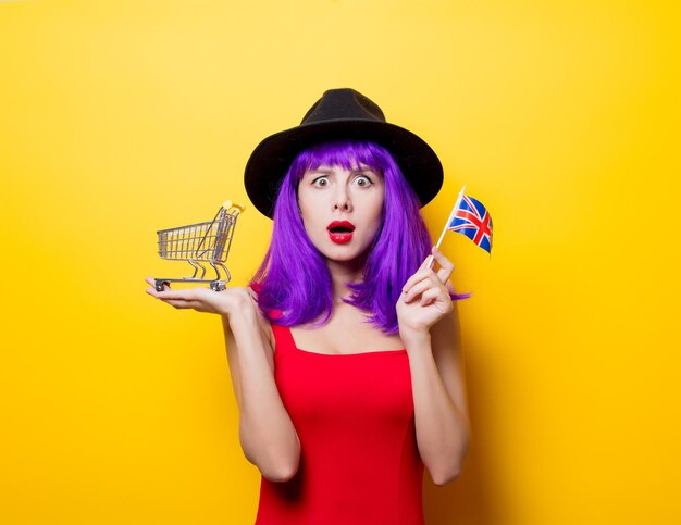 Portret van jonge stijl hipster meisje met paars kapsel met winkelwagen en Groot-Brittannië vlag in de hand op gele achtergrond