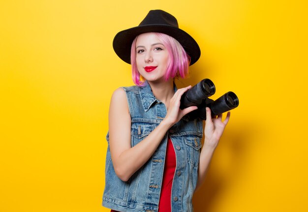 Portret van jonge stijl hipster meisje met roze kapsel met verrekijker op gele achtergrond