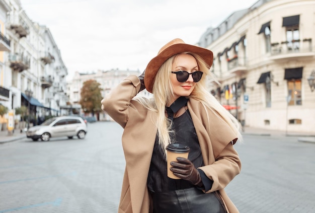 Portret van jonge stijlvolle blonde vrouw in herfstjas, zonnebril, handschoenen en vilten hoed met koffiekopje in Europese stad. Mooi aantrekkelijk meisje