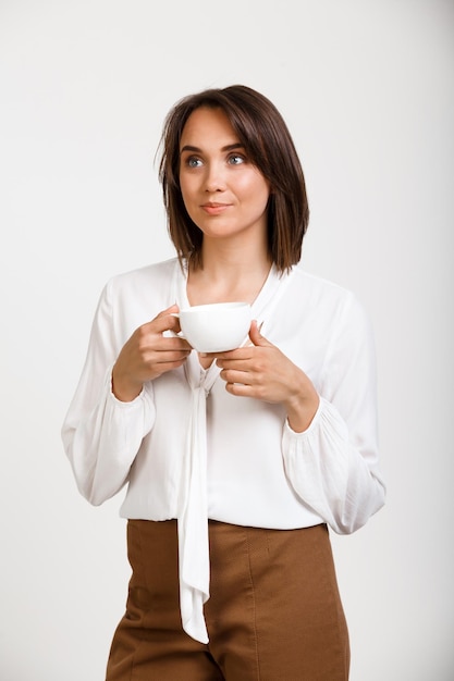 Portret van jonge succesvolle zakenvrouw, glimlachend, kopje koffie houdend, over witte muur.