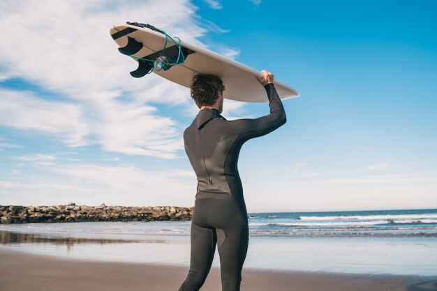 Portret van jonge surfer bij het strand dat zijn surfplank steunt en een zwart surfend kostuum draagt. sport en watersport concept.