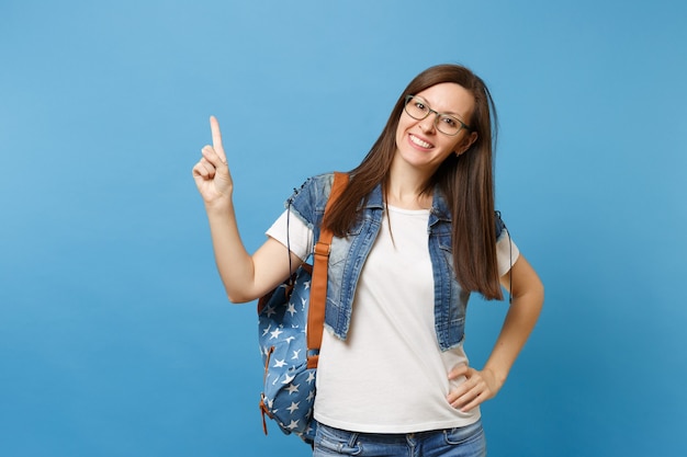 Portret van jonge vrolijke studente in denim kleding, bril met rugzak wijzende wijsvinger omhoog op kopie ruimte geïsoleerd op blauwe achtergrond. Onderwijs in het concept van de middelbare schooluniversiteit.