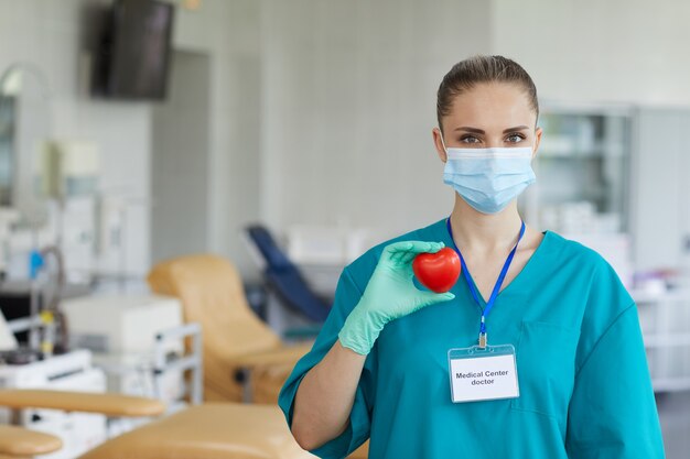 Portret van jonge vrouwelijke arts in masker en in eenvormige hartvorm in haar handen en