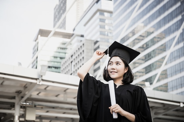 Portret van jonge vrouwelijke gediplomeerden in vierkante academische GLB die gelukkig holdingsdiploma glimlachen tegen de bouw.