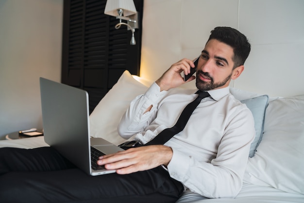 Portret van jonge zakenman liggend op bed en praten aan de telefoon tijdens het werken op zijn laptop in de hotelkamer. zakelijke reizen concept.