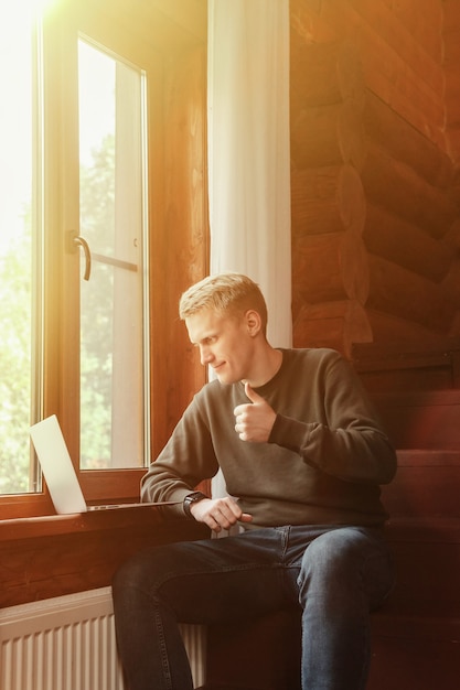 Portret van jonge zakenman met laptop bij raam van woonkamer in landhuis. Workaholic man in huis casual kleding aan het werk op vakantie. Creatieve inspiratie en startende onderneming. Ruimte kopiëren