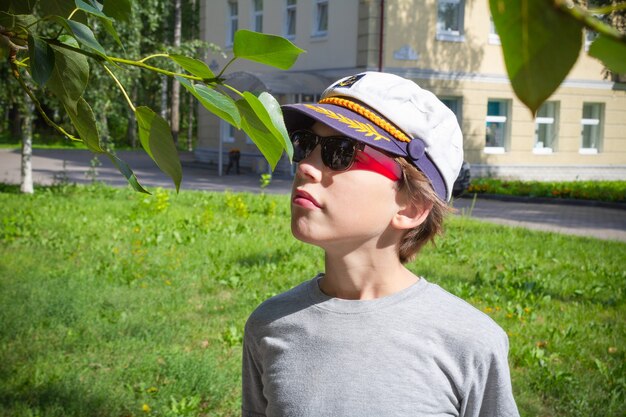 Portret van jongen in witte pet, zonnebril en grijs t-shirt snuivende tak met groene bladeren op zonovergoten groene stadsstraat