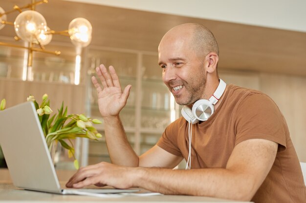 Portret van kale volwassen man glimlachend en zwaaien naar de camera tijdens het spreken door videochat via laptop in modern interieur