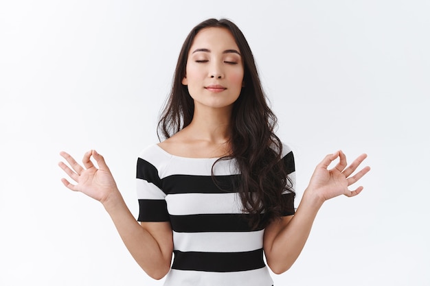 Portret van kalme en opgeluchte Oost-Aziatische vrouw houdt de geest gezond, ontspant met yoga of mediteert, diep adem en sluit de ogen, houd handen zijwaarts met mudra zen-tekens, witte achtergrond