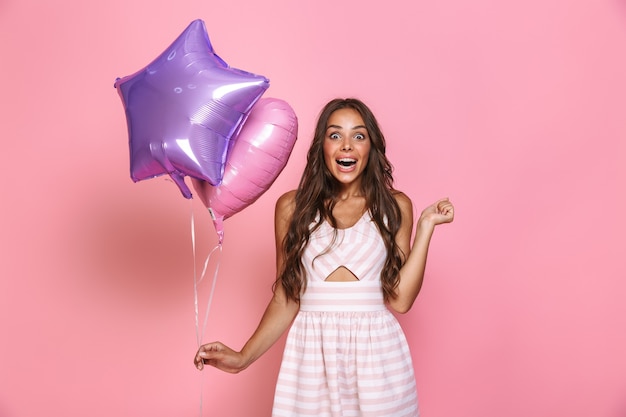 Portret van Kaukasisch blij meisje 20s met lang haar gekleed in een jurk glimlachend en met twee ballonnen, geïsoleerd over roze muur