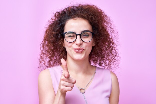 Portret van Kaukasisch krullend lachend meisje wijst vinger naar camera en knipoogt, kiest je concept geïsoleerd in roze studio met kopie ruimte